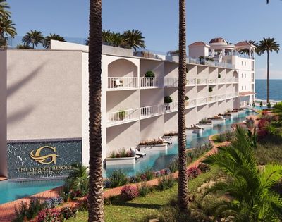 A modern white resort building with multiple balconies, swimming pools, and lush tropical landscaping, situated on a sandy beach with a calm blue ocean in the background.