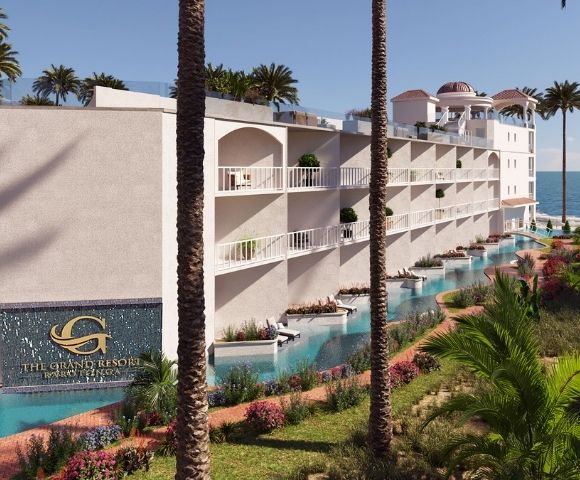 A modern white resort building with multiple balconies, swimming pools, and lush tropical landscaping, situated on a sandy beach with a calm blue ocean in the background.