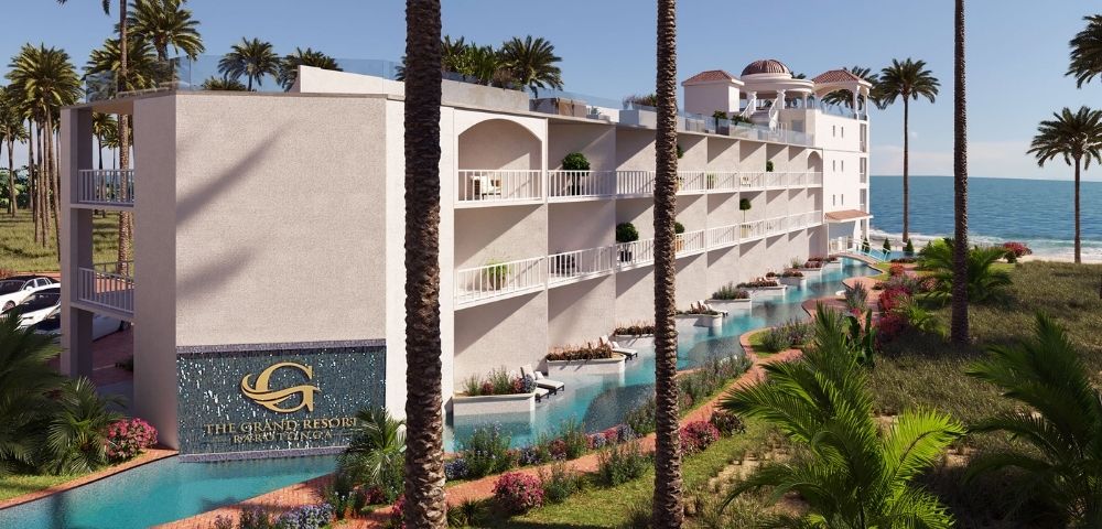 A modern white resort building with multiple balconies, swimming pools, and lush tropical landscaping, situated on a sandy beach with a calm blue ocean in the background.