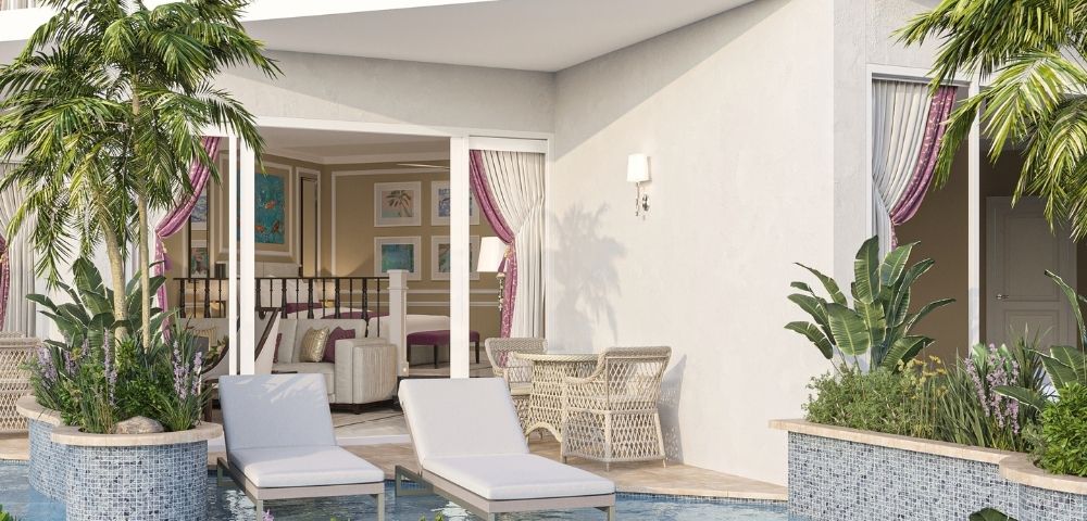 A luxurious hotel suite with a private patio featuring lounge chairs, a table with chairs, lush tropical plants, and a pool.