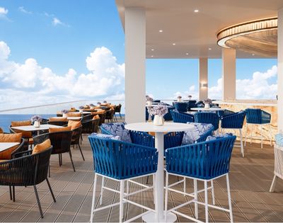 An upscale rooftop bar and dining area overlooking the ocean, featuring blue wicker chairs with patterned cushions, tables with orange-cushioned chairs, a long bar with stools, and a circular wooden ceiling fixture.