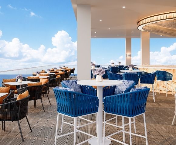 An upscale rooftop bar and dining area overlooking the ocean, featuring blue wicker chairs with patterned cushions, tables with orange-cushioned chairs, a long bar with stools, and a circular wooden ceiling fixture.