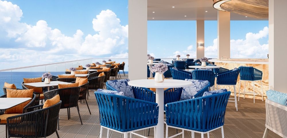 An upscale rooftop bar and dining area overlooking the ocean, featuring blue wicker chairs with patterned cushions, tables with orange-cushioned chairs, a long bar with stools, and a circular wooden ceiling fixture.