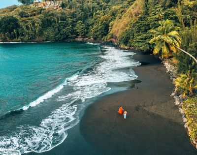 Aerial view of a black sand beach with turquoise water and lush green cliffs, two people walking on the sand.