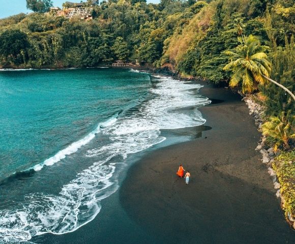 Aerial view of a black sand beach with turquoise water and lush green cliffs, two people walking on the sand.