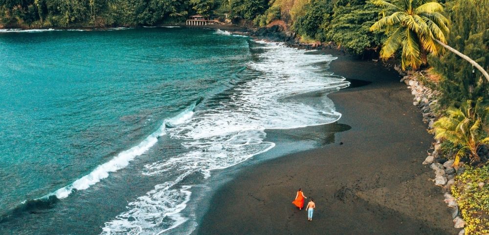 Aerial view of a black sand beach with turquoise water and lush green cliffs, two people walking on the sand.