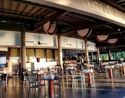 An outdoor dining area with many tables and chairs set up under a thatched roof.