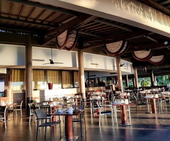 An outdoor dining area with many tables and chairs set up under a thatched roof.
