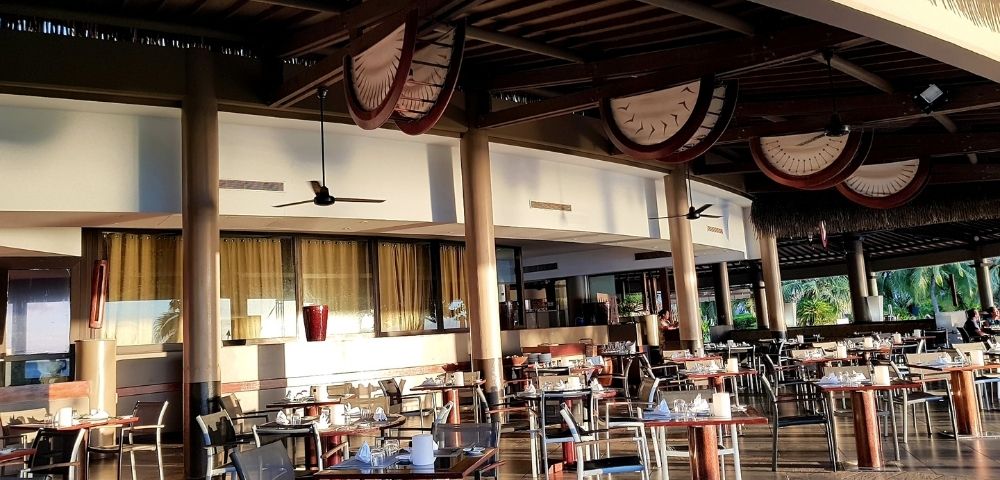An outdoor dining area with many tables and chairs set up under a thatched roof.