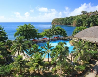 Resort swimming pool surrounded by palm trees with a view of the ocean and a lush green coastline.