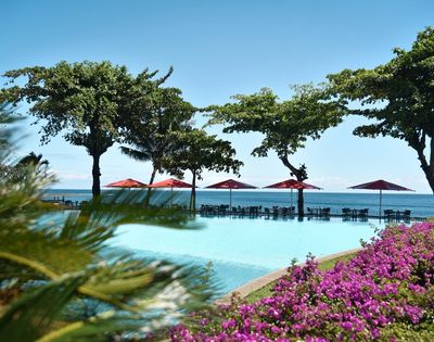 An empty infinity pool reflects the clear blue sky, with tables and red umbrellas set up on the deck overlooking the ocean. Lush green trees line the edge of the pool, and vibrant purple flowers bloom in the foreground.