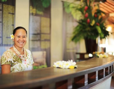 A smiling person wearing a floral shirt, a lei, and a white flower in their hair stands behind a reception desk decorated with leis.