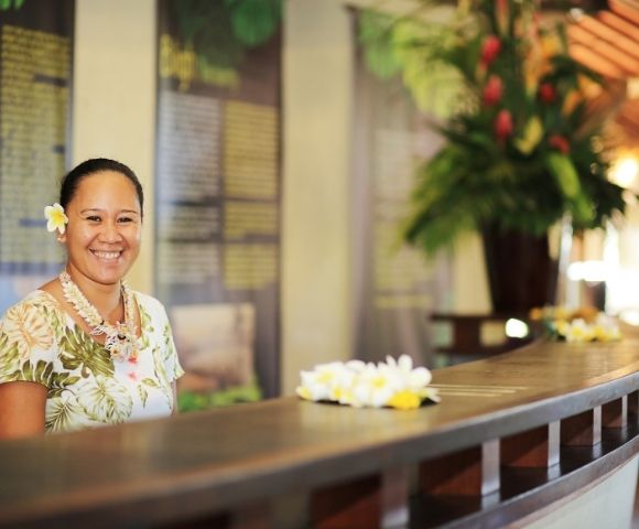 A smiling person wearing a floral shirt, a lei, and a white flower in their hair stands behind a reception desk decorated with leis.