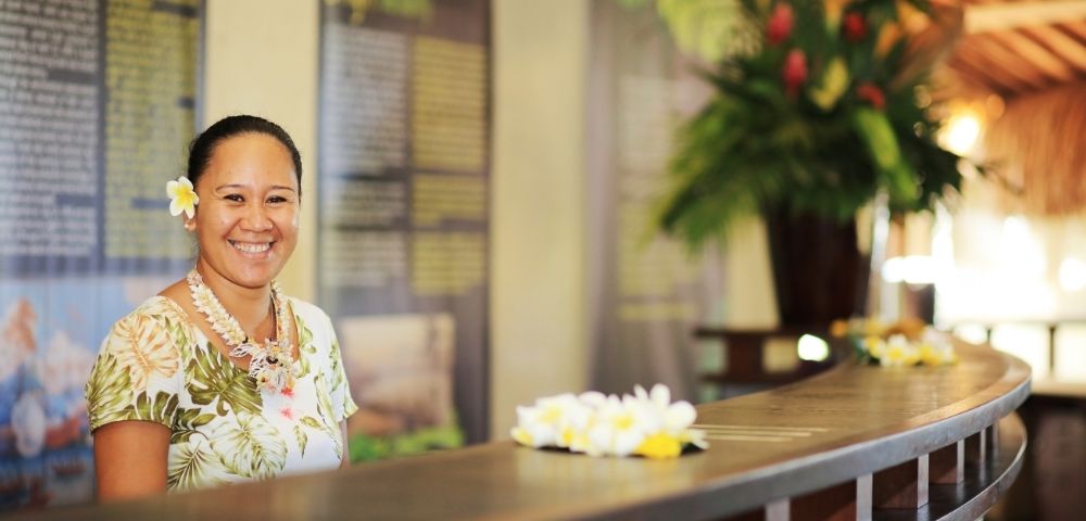 A smiling person wearing a floral shirt, a lei, and a white flower in their hair stands behind a reception desk decorated with leis.