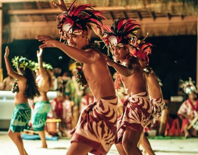 Dancers in traditional headdresses and patterned wraps perform outdoors at dusk.