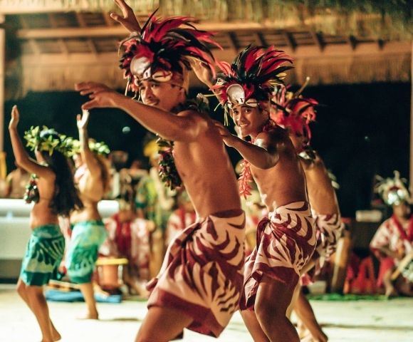 Dancers in traditional headdresses and patterned wraps perform outdoors at dusk.