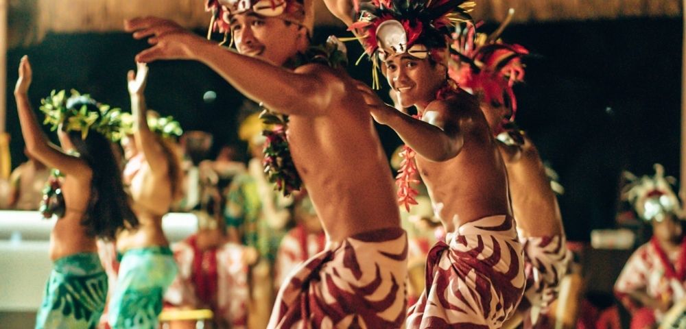Dancers in traditional headdresses and patterned wraps perform outdoors at dusk.