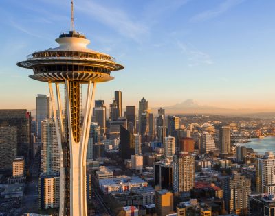 Panoramic aerial view of the Seattle skyline at sunset, featuring the Space Needle prominently in the foreground, with numerous skyscrapers, Elliott Bay, industrial areas, and a distant snow-capped mountain under a clear, warm-toned sky.