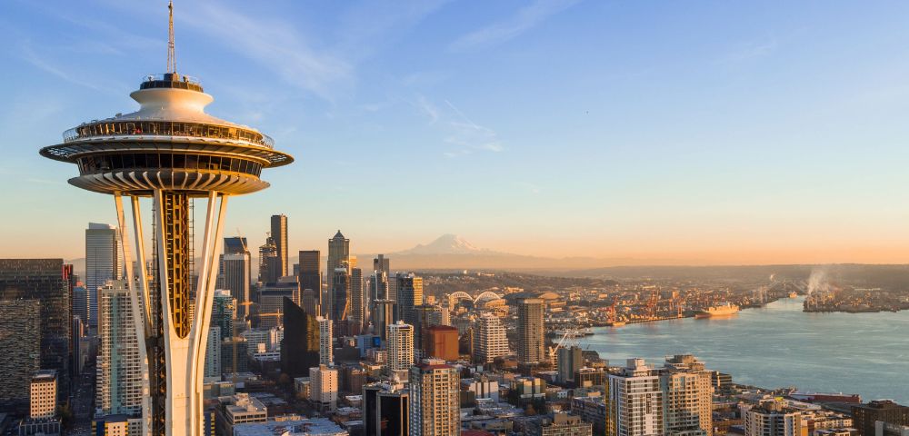 Panoramic aerial view of the Seattle skyline at sunset, featuring the Space Needle prominently in the foreground, with numerous skyscrapers, Elliott Bay, industrial areas, and a distant snow-capped mountain under a clear, warm-toned sky.