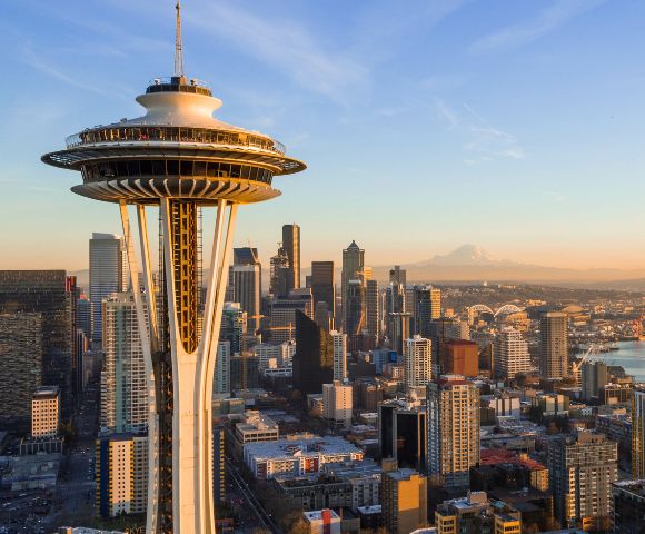 Panoramic aerial view of the Seattle skyline at sunset, featuring the Space Needle prominently in the foreground, with numerous skyscrapers, Elliott Bay, industrial areas, and a distant snow-capped mountain under a clear, warm-toned sky.