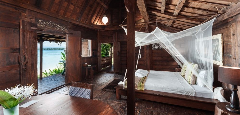 Rustic wooden bedroom with a mosquito net draped over a large bed, opening to a view of the ocean and lush greenery.