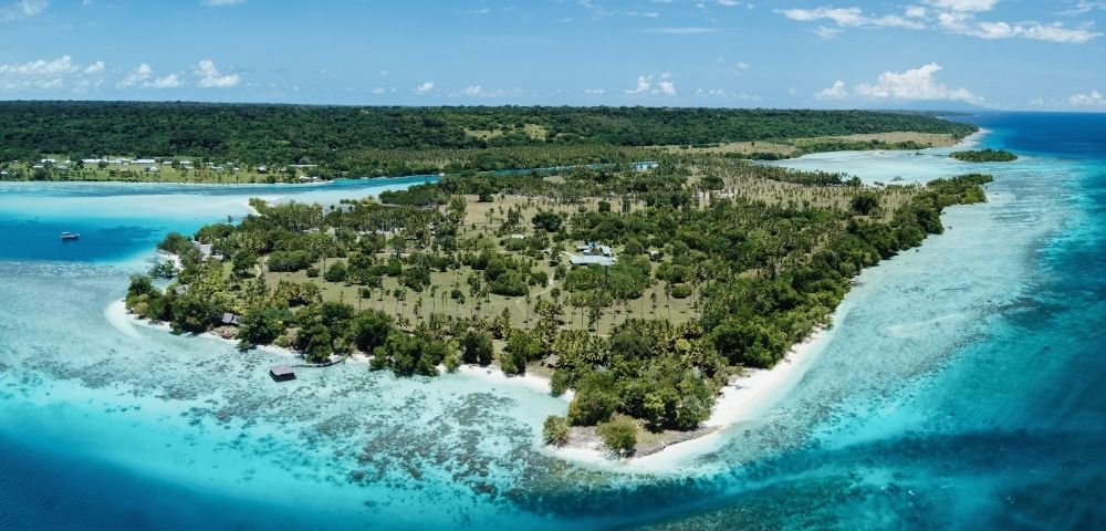 Aerial view of a lush, tropical island with white sand beaches, surrounded by clear turquoise waters and coral reefs. Dense green vegetation covers the island's interior, with scattered palm trees and a few small buildings visible. The ocean exhibits varying shades of blue, indicating depth and coral formations.