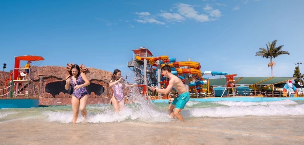 Three young people play in the shallow waves of a water park on a sunny day, with colorful water slides in the background.
