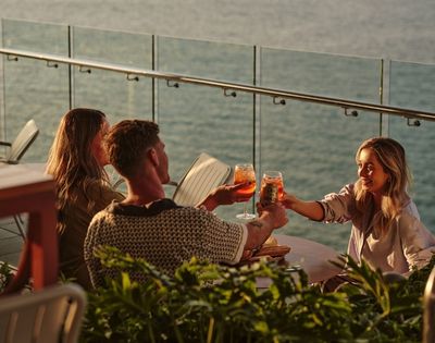 Three people are toasting drinks on a patio overlooking the water at sunset.