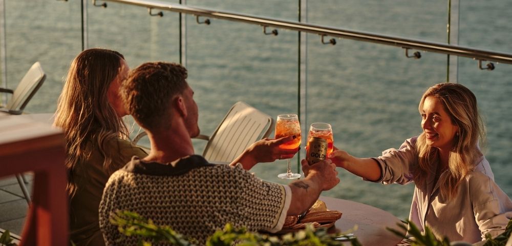 Three people are toasting drinks on a patio overlooking the water at sunset.