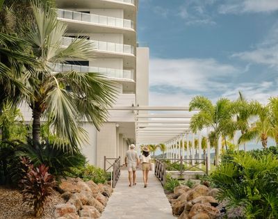A couple strolls through a sunny tropical resort walkway, framed by palm trees, lush greenery, white balconies, and a calm, inviting atmosphere.