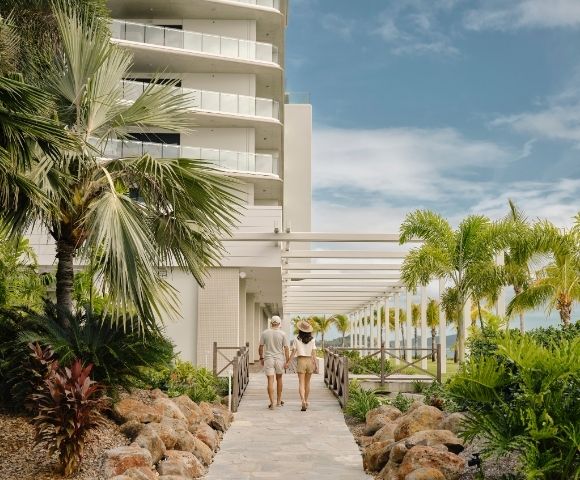 A couple strolls through a sunny tropical resort walkway, framed by palm trees, lush greenery, white balconies, and a calm, inviting atmosphere.
