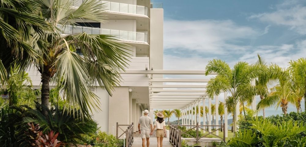 A couple strolls through a sunny tropical resort walkway, framed by palm trees, lush greenery, white balconies, and a calm, inviting atmosphere.