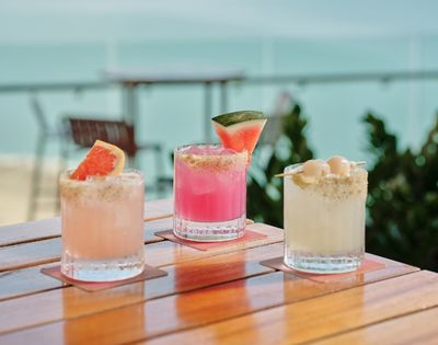 Three colorful summer cocktails sit on a wooden patio table: peach, pink watermelon, and creamy coconut drinks with bright garnishes.
