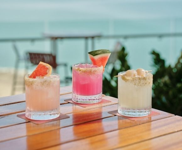 Three colorful summer cocktails sit on a wooden patio table: peach, pink watermelon, and creamy coconut drinks with bright garnishes.