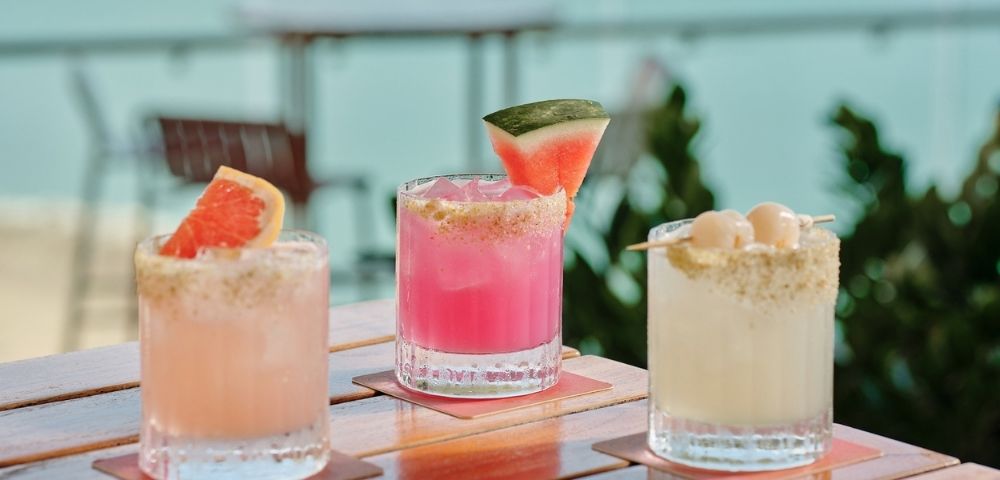 Three colorful summer cocktails sit on a wooden patio table: peach, pink watermelon, and creamy coconut drinks with bright garnishes.