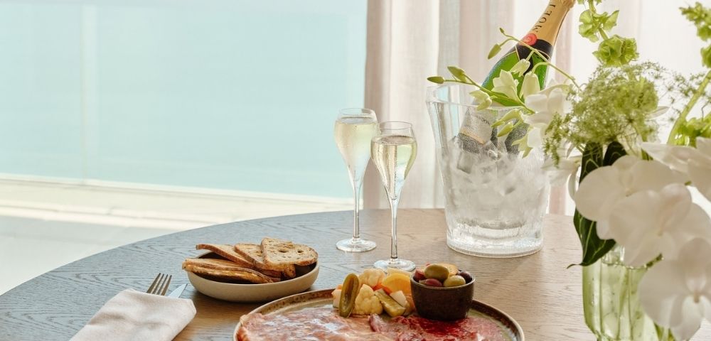 Elegant seaside brunch table with salmon platter, toast and fruit, champagne flutes, flowers, and an ice bucket, creating a serene mood.