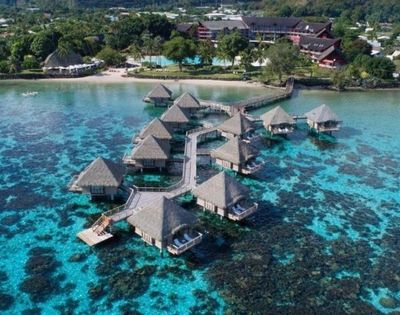 Aerial view of turquoise tropical waters, coral reefs, and thatched overwater bungalows connected by walkways near a lush island resort.