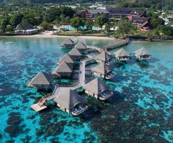 Aerial view of turquoise tropical waters, coral reefs, and thatched overwater bungalows connected by walkways near a lush island resort.