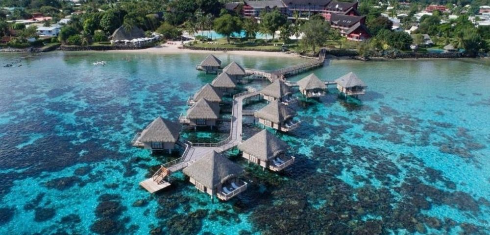 Aerial view of turquoise tropical waters, coral reefs, and thatched overwater bungalows connected by walkways near a lush island resort.