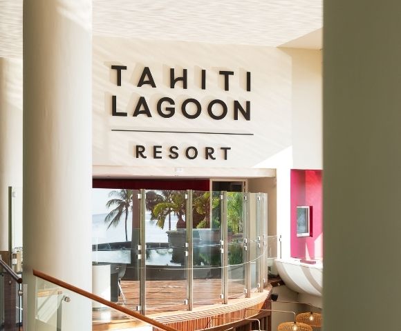 Bright Tahiti Lagoon Resort lobby with bold wall signage, glass doors, tropical palms, and ocean views creating a welcoming island atmosphere.