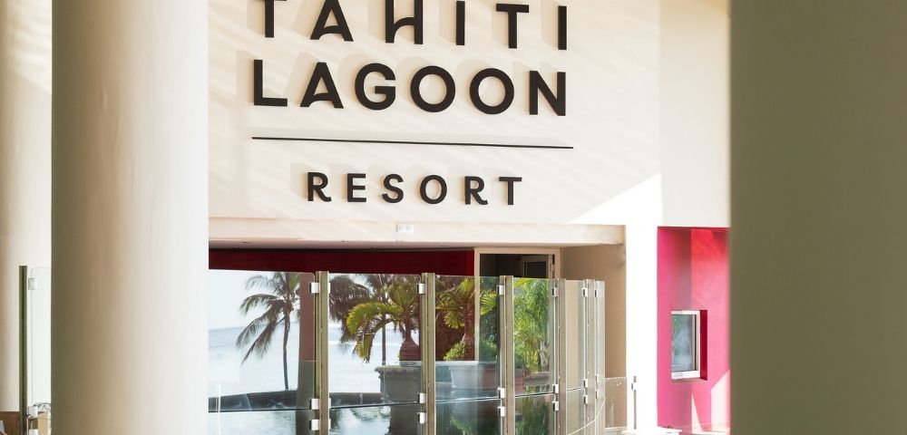 Bright Tahiti Lagoon Resort lobby with bold wall signage, glass doors, tropical palms, and ocean views creating a welcoming island atmosphere.