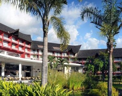 Tropical resort with red-and-white buildings, thatched roofs, palm trees, lush gardens, and a bright blue sky creating a relaxed mood.