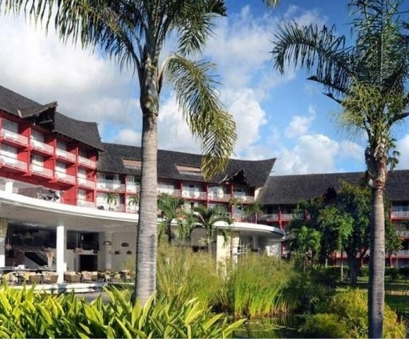 Tropical resort with red-and-white buildings, thatched roofs, palm trees, lush gardens, and a bright blue sky creating a relaxed mood.