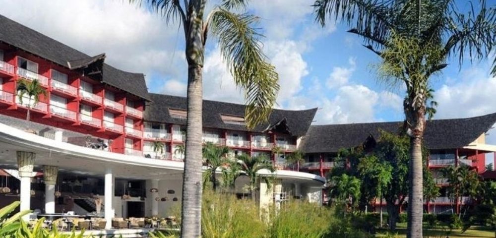 Tropical resort with red-and-white buildings, thatched roofs, palm trees, lush gardens, and a bright blue sky creating a relaxed mood.