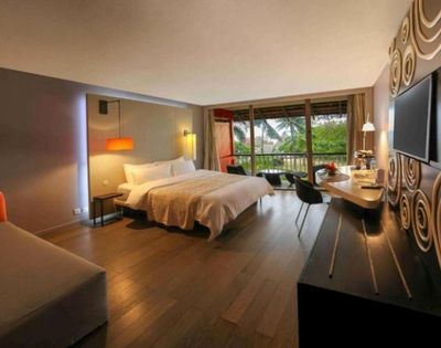 Modern luxury hotel room with king bed, sofa, desk, wall-mounted TV, and balcony overlooking tropical greenery; warm, stylish, inviting.