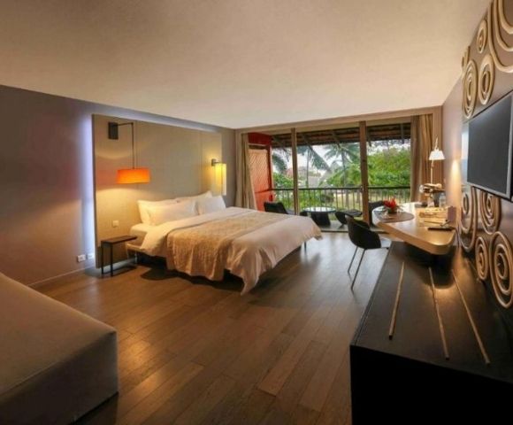 Modern luxury hotel room with king bed, sofa, desk, wall-mounted TV, and balcony overlooking tropical greenery; warm, stylish, inviting.