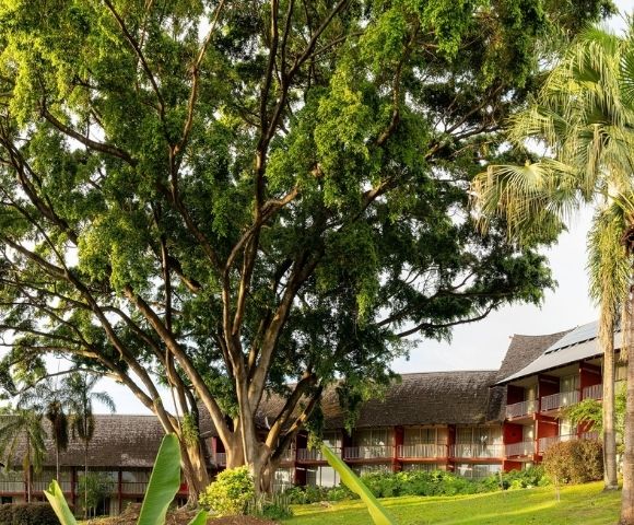 Lush tropical resort with red-roofed buildings, tall palms, flowering gardens, and a broad shade tree creating a peaceful, inviting atmosphere.