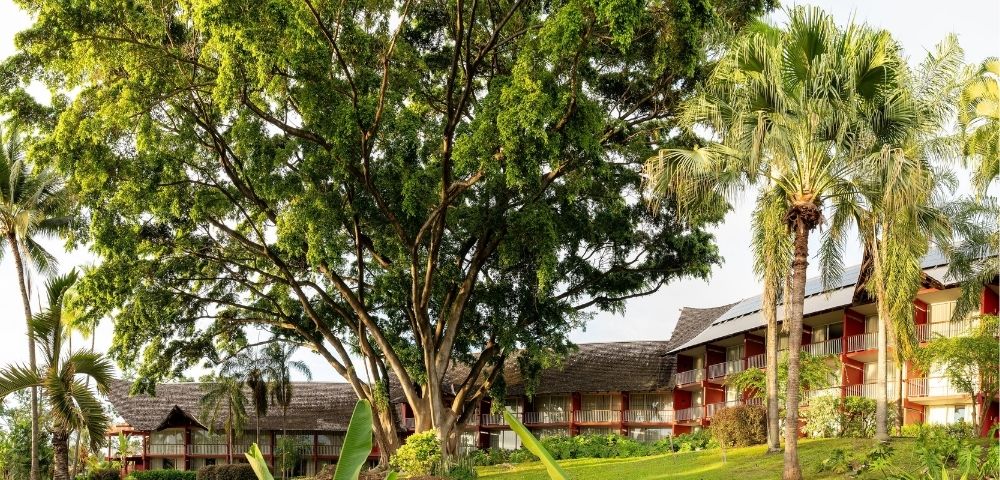 Lush tropical resort with red-roofed buildings, tall palms, flowering gardens, and a broad shade tree creating a peaceful, inviting atmosphere.