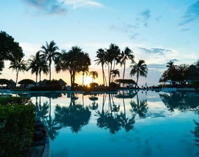 Tropical resort pool at sunset, with tall palm trees, thatched huts, and a glowing sky reflected in calm blue water; peaceful mood.