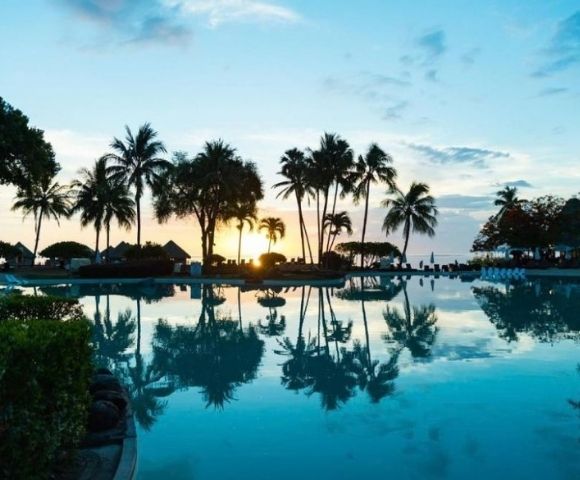 Tropical resort pool at sunset, with tall palm trees, thatched huts, and a glowing sky reflected in calm blue water; peaceful mood.